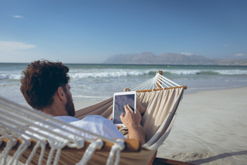 Man using digital tablet while lying on hammock at beach 
