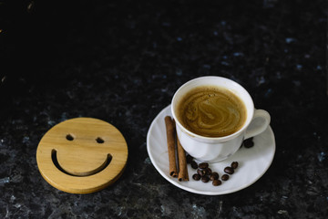 Wooden smile on a dark black texture background. On the edge you can find a cup of coffee with espresso, on the saucer. No people, no body.