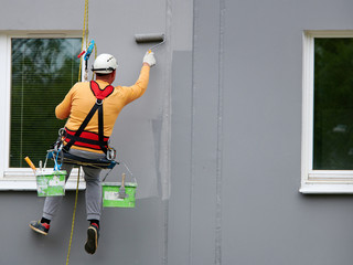 Worker hanging on rope and paints building wall with roller. Painter hanging on cable with paint...