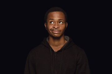 Close up portrait of young african-american man isolated on black studio background. Real emotions of male model. Standing and dreaming, has a doubt. Facial expression, human nature and emotions