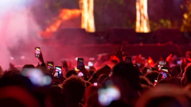 People are jumping with their hands up, holding their smartphones and watching a concert. All is illuminated with stage light. 4K