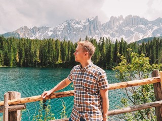 A young blond man in a plaid shirt and with a backpack stands near the mountain lake Carezza, a romantic beautiful place, azure pure water. Mountain summer landscape in the Dolomites in Northern Italy