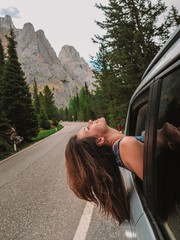 Brunette girl with long hair leans out of the car window against the background of the dolomite Mountains