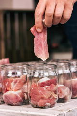 Hand is putting a piece of meat in a jar. Food preservation. Preparation of homemade pork stew. Process close up. Vertical.