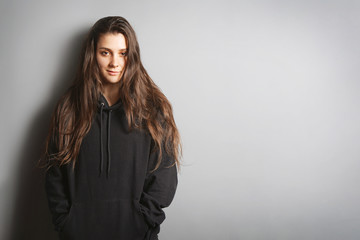 cool young woman with long brunette hair wearing black hoodie sweater leaning against wall with her hands in pockets - gray background with copy space