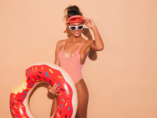 Young beautiful sexy smiling hipster woman in sunglasses.Girl in summer swimwear bathing suit with donut lilo inflatable mattress.Positive female going crazy.Near beige wall in transparent visor cap - obrazy, fototapety, plakaty