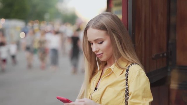 A blond girl with straight hair and yellow shirt enjoying some music on her phone in the street