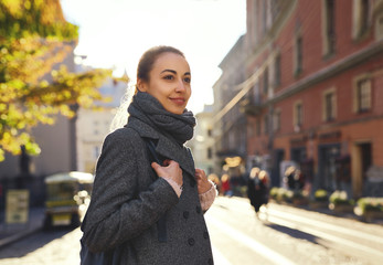 Young attractive woman in a warm scarf and gray coat walking city street. Stylish smiling girl posing at camera, enjoying a sunny autumn day.