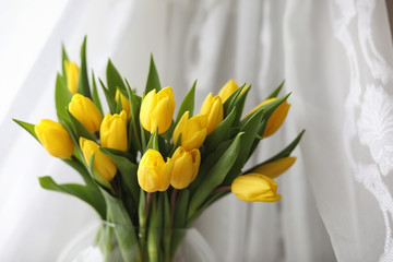 A bouquet of yellow tulips in a vase on the windowsill. A gift to a woman's day from yellow tulip flowers. Beautiful yellow flowers in a vase by window.