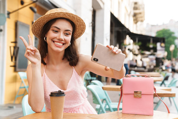 Image of charming brunette woman smiling and taking selfie photo cellphone while sitting in street summer cafe