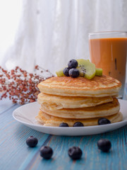 Stack of pancakes with topping, kiwi and blueberry.placed in a white plate on a blue wooden table.Eat with milk tea in the glass.