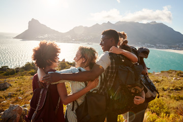 Millennial friends on a hiking trip embrace at the summit to celebrate their climb, back view - obrazy, fototapety, plakaty