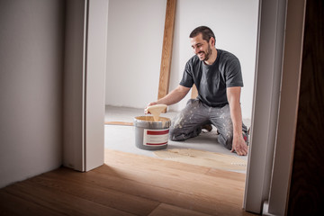 Handwerker verlegt Parkettboden und streicht den Estrich mit Kleber ein und schlägt die Dielen mit...