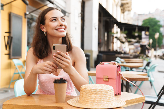 Image of happy brunette woman smiling and holding cellphone while sitting in street summer cafe