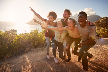 Five millennial friends on a road trip have fun posing for photos on a coastal path, full length