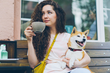 Attractive girl is drinking tea outdoors in cafe and patting cute shiba inu dog sitting on bench in the street enjoying day out. People, pets and lifestyle concept.