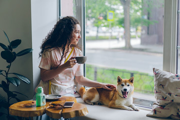 Happy student pretty girl is stroking adorable shiba inu dog sitting in cafe on window sill with...