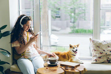 Pretty girl is listening to music with headphones and using smartphone sitting on window sill in...