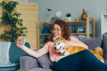 Happy young girl is taking silfie with lovable dog using smartphone camera having fun sitting on couch holding device. Youth and domestic animals concept.