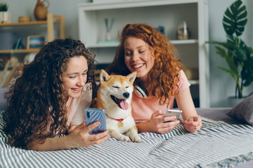 Beautiful curly-haired young women sisters talking using smartphones and caressing pedigree dog lying on bed in apartment. People and gadgets concept. - Powered by Adobe