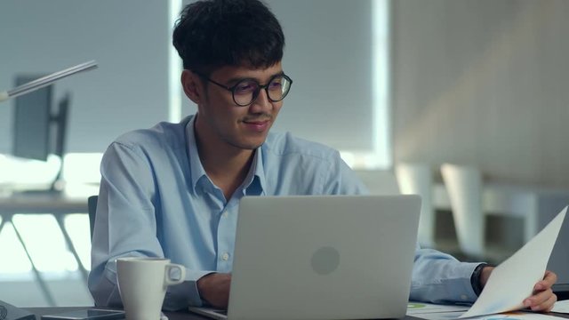 Asian creative man working on laptop. Young professional business male using computer looking information paper, business situation, startup in Loft office concept.