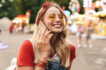 Smiling pleased young cheery blonde woman in amusement park talking by mobile phone.