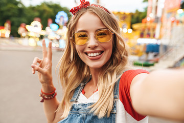 Image of young blonde woman showing peace sign and taking selfie photo at amusement park