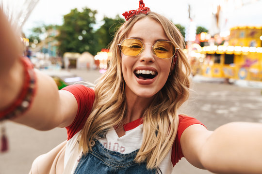 Image of adorable blonde woman laughing and taking selfie photo at amusement park