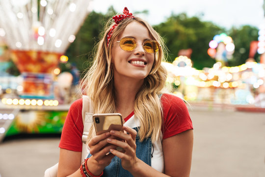 Image of cute blonde woman holding cellphone and walking in front of colorful carousel at amusement park
