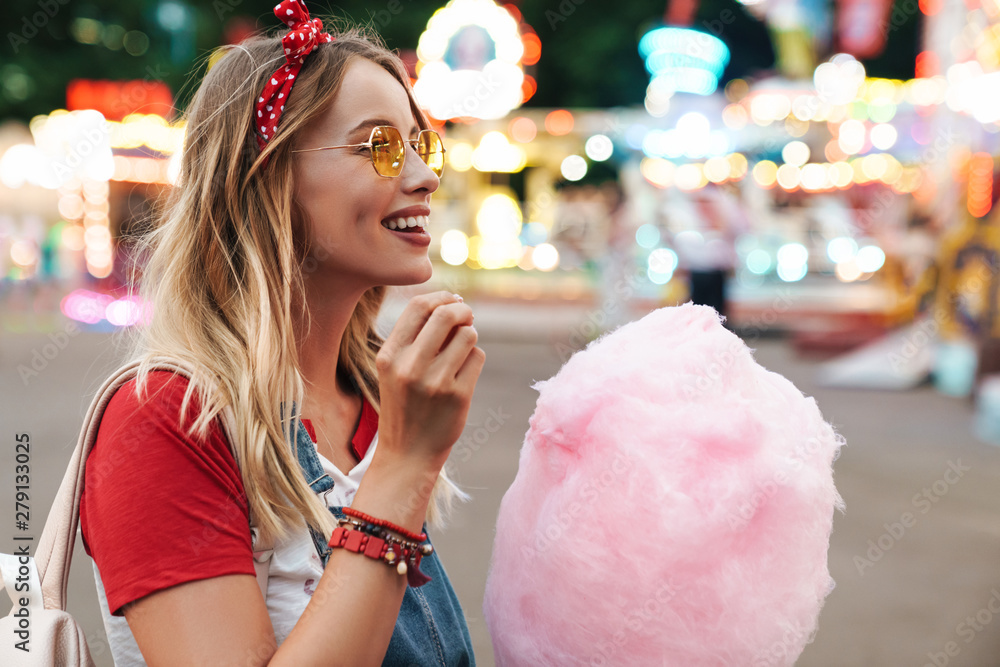 Wall mural Image of blonde beautiful woman eating sweet cotton candy while walking in amusement park