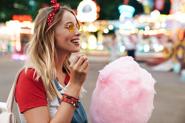 Image of blonde beautiful woman eating sweet cotton candy while walking in amusement park