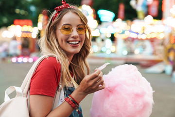 Image of positive blonde woman eating sweet cotton candy while walking in amusement park