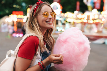 Image of cheery blonde woman eating sweet cotton candy while walking in amusement park