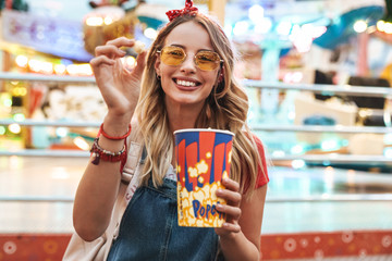 Image of young blonde woman smiling and holding popcorn while walking in amusement park