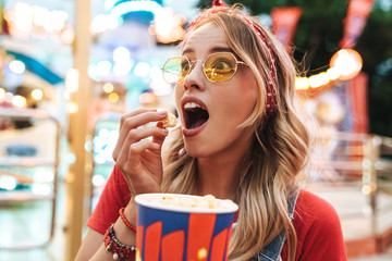 Image of surprised blonde woman rejoicing and holding popcorn while walking in amusement park