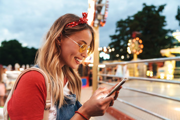 Image of cute blonde woman smiling and holding cellphone in front of colorful carousel at amusement...