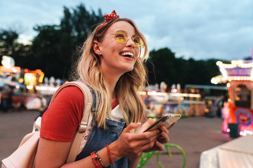 Image of adorable blonde woman smiling and holding cellphone in front of colorful carousel at...