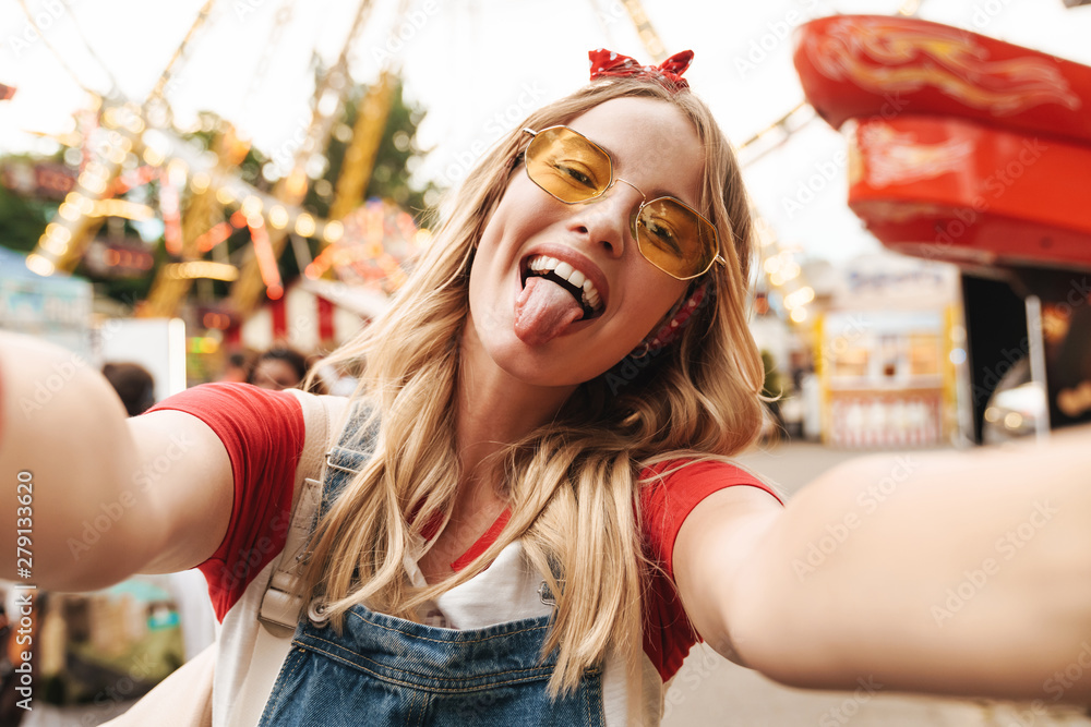 Wall mural Image of positive blonde woman sticking out her tongue and taking selfie photo at amusement park