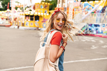 Image of beautiful blonde woman smiling and walking in front of colorful carousel at amusement park