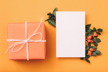 Holiday greeting set. White mockup paper, peach gift box with rose hip decor on orange background....