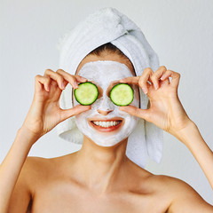 Beautiful young woman with facial mask on her face holding slices of cucumber. Skin care and treatment, spa and cosmetology concept.