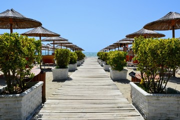 Obraz premium Wooden walkway leading to the beach and the coast between flower pots and beach umbrellas