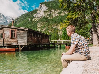 The blonde man in a plaid shirt sits on a background of Lake Lago di Braies in the Dolomites, South Tyrol, Italy. Pier with romantic old wooden rowing boats on the lake. Amazing view of lake Lago di B