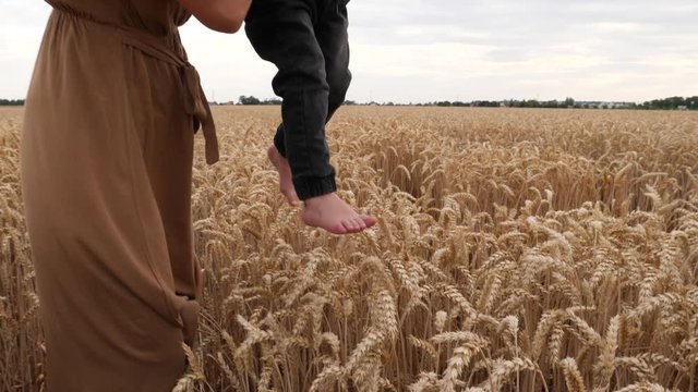 Young mother holds little boy son as child slightly touching wheat ears with barefoot legs feet toes on countryside nature field