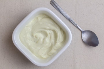 Bowl of creamy vanilla yogurt with spoon on linen table cloth surface - top view of vanilla yoghurt in square ceramic dish