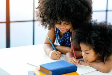 African girls as older and younger sister write or draw something on white paper near the book in front of glass windows with day light.