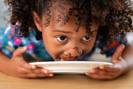 Close Up Of Little Girl Licking Chocolate Off Plate With Messy Face