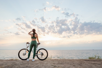 Fototapeta premium Beautiful young brunette woman resting after bicycle ride at sunrise. Model listening to music with wireless earbuds in the morning.