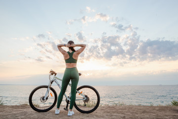 Fototapeta premium Beautiful young brunette woman resting after bicycle ride at sunrise. Model listening to music with wireless earbuds in the morning.