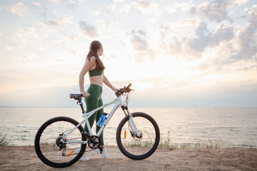 Obraz premium Beautiful young brunette woman resting after bicycle ride at sunrise. Model listening to music with wireless earbuds in the morning.